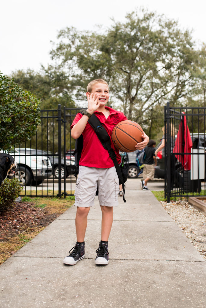 Male student stands at school gate with basketball and waves